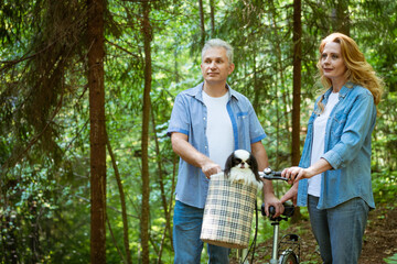 an adult couple in denim clothes on a walk in the woods with a dog that sits in the basket of a bicycle.