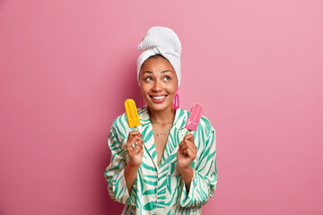 Domestic style and summer dessert concept. Positive dark skinned woman smiles happily holds two delicious ice creams on sticks wears silk pajama wrapped bath towel on head has happy mood poses indoor