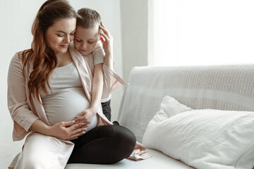 Little boy hugs the belly of a pregnant mom at home on the couch.