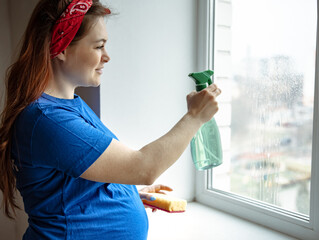 Young pregnant woman cleans windows with spray and sponge.
