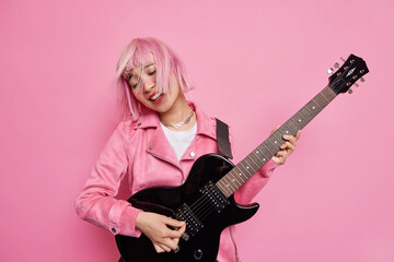 Cool stylish female rocker plays famous song on electric guitar tilts head has pink hair floating on wind demonstrates her talent wears jacket isolated over rosy studio background. Musical instruments