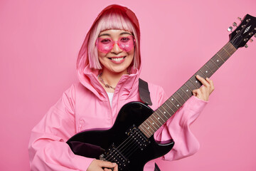 Obraz premium People entertainment and music concept. Cheerful fashionable female guitarist wers jacket with hood plays electric guitar prepares for hardrock concert or rock festival isolated on pink wall