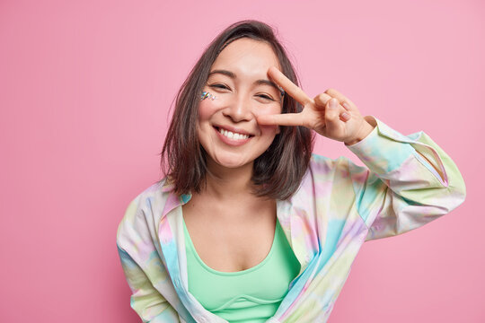 Cute positive Asian woman with dark hair shows v sign peace gesture looks happily at camera dressed in casual shirt enjoys good day isolated over pink background expresses carefree emotions.