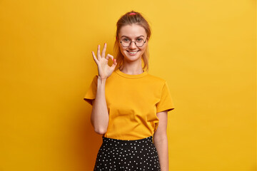 Pleased young European woman makes okay gesture shows excellent job says alright satisfied with...