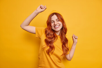Fototapeta premium Studio shot of carefree redhead woman raises hand dances and smiles cheerfully has fun wears hat and casual t shirt isolated over vivid yellow background partying in good mood. Monochrome shot.
