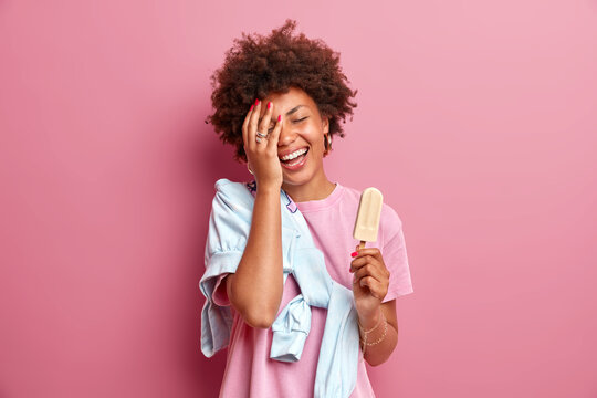 Smiling teenage girl makes face palm feels glad wears casual t shirt with jumper enjoys summer time eats delicious ice cream on stick has good mood isolated over pink background. Fun and sweets