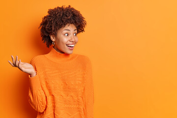 Horizontal shot of happy excited woman raises palm notices something unexpected and surprising smiles positively wears casual jumper isolated over vivid orange background blank copy space away