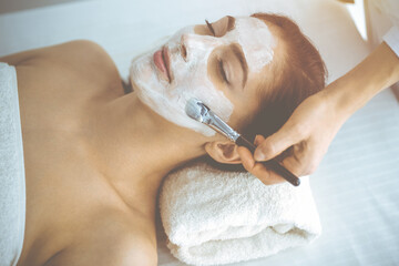 Beautiful brunette woman enjoying applying cosmetic mask with closed eyes. Relaxing treatment in medicine and spa concepts