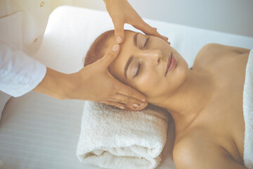 Obraz na płótnie Canvas Beautiful brunette woman enjoying facial massage with closed eyes in sunny spa center. Relaxing treatment and cosmetic medicine concepts