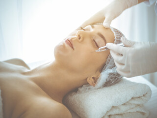 Beautician doing beauty procedure with syringe to face of young brunette woman in sunny clinic. Cosmetic medicine and surgery, beauty injections