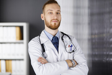 Red-bearded doctor standing straight in clinic near his working place. Portrait of physician. Medicine, healthcare