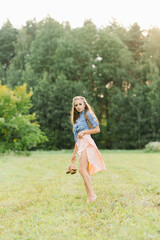A beautiful young woman holds summer shoes in her hands and walks barefoot on the grass on a summer day