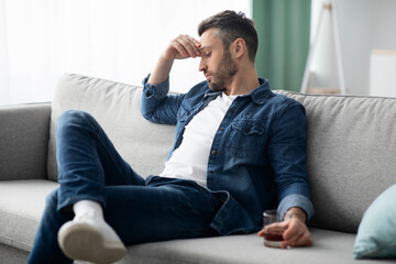 Depressed middle-aged man sitting on couch with glass of alcohol