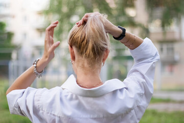 The blonde girl stands with her back to the camera and pulls her hair into a ponytail.