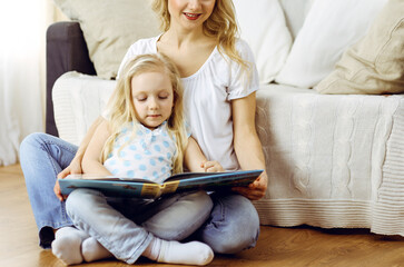 Happy family. Blonde young mother reading a book to her cute daughter while sitting at wooden floor. Motherhood concept