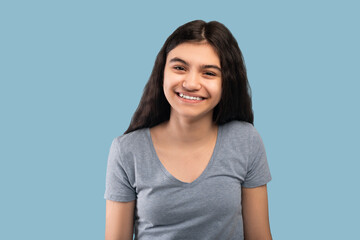 Beautiful Indian teenage girl with dark long hair smiling and looking at camera on blue studio...