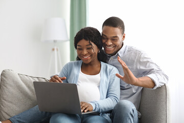 Cheerful pregnant black couple having video call, using laptop