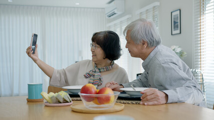 Young senior asia citizen couple hold mobile phone selfie videocall online to family at home apartment morning on dining table in older people with digital technology virtual talk conference concept.