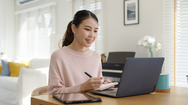 Candid young attractive beautiful asian businesswoman work remotely from home or learning online sitting smiling in cozy workspace at home feeling happy in quarantine social distance concept.