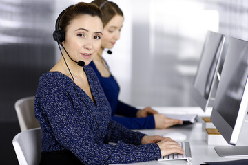 Two busineswomen have conversations with the clients by headsets, while sitting at the desk in a modern office. Diverse people group in a call center. Telemarketing and customer service