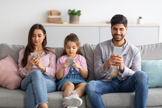 Gadget Addiction Concept. Young Arab Family Of Three Holding And Using Smartphones While Sitting On The Sofa In Living Room