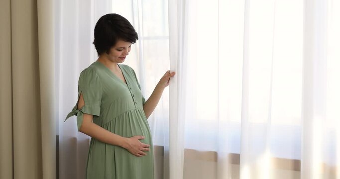 Young smiling pregnant woman in dress strokes her big belly while stand at home near window touch curtain looking outside. Unborn child anticipation, happy maternity, motherhood, child bearing concept