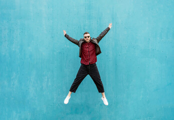 Fototapeta na wymiar Positive man in casual clothes jumping outdoor with open arms against a blue wall background - Trendy guy having fun jumping with open arms - Motivation, braving and victory concept - Copy space