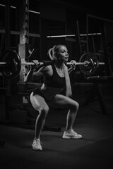 Fototapeta na wymiar A fit woman with blonde hair is squatting with a barbell near the squat rack in a gym. A girl is doing a leg workout.