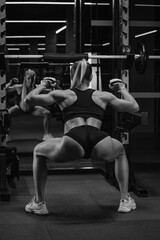 Fototapeta na wymiar A photo from behind of a sporty woman with blonde hair who is warming up before squatting with a barbell near the squat rack in a gym. A girl is doing a leg workout.