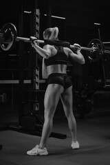 Fototapeta na wymiar A photo from behind of a sporty woman with blonde hair who is started squatting with a barbell near the squat rack in a gym. A girl is doing a leg workout.