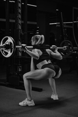 Fototapeta na wymiar A photo from behind of a fit woman with blonde hair who is squatting with a barbell near the squat rack in a gym. A girl is doing a leg workout.
