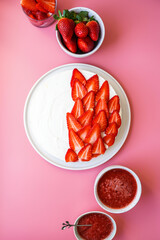 A top view on a white cheesecake with slices of strawberries, fresh strawberries and jam on a pink surface