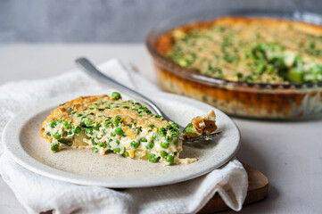 A slice of savoury pie with peas on beige plate on light-colored napkin and a whole shepherd's pie on the background