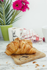 A sunny easter breakfast meal consisting of a butter croissant on a wooden board and a cup of coffee and several magenta gerberas in a green vase behind
