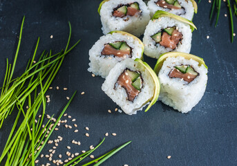 Sushi roll with salmon and avocado on black surface surrounded by greens, salad leaves and sesame seeds