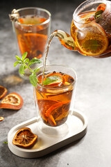 A transparent tea pot filled with black tea, oranges and mint on a grey surface pouring tea into a glass
