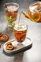 A transparent tea pot filled with black tea, oranges and mint on a grey surface pouring tea into a glass
