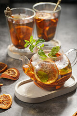 A transparent tea pot filled with black tea, oranges and mint on a grey surface and two glasses in the background