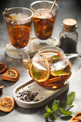 A transparent tea pot filled with black tea, oranges and mint on a grey surface and two glasses in the background