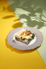 A piece of quiche, french pie filled with spinach, feta cheese and dried tomatoes on a romantic plate on a double colored (yellow and green) background in hard light