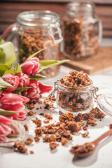 Mason jar with granola breakfast cereal with nuts. wooden spoon with scattered granola and a bouquet of tulips and two jars with granola on small wooden board in the back
