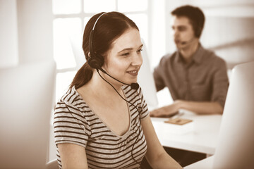 Casual dressed young woman using headset and computer while talking with customers online. Group of operators at work. Call center, business concept