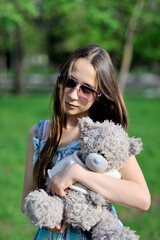 A charming teenage girl in sunglasses stands and looks at the camera in a park on a sunny summer day with a soft toy in her hands. Close-up. Fashion portrait. Vertical orientation.