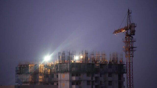 night evening blue hour shot aerial drone view of a construction site on a skyscraper rooftop with rebar sticking out and workers labourers moving around in helmets while a huge crane moves and lights