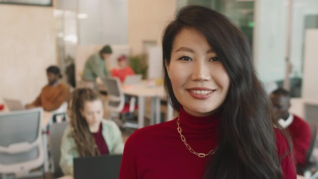 Portrait PAN with close up of happy young Asian businesswoman smiling for camera in open-plan office