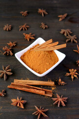 Bowl of ground cinnamon powder and sticks with star anise scattered on black wooden table