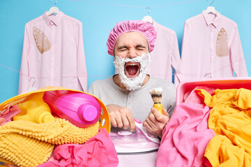 Emotional man keeps mouth opened shouts loudly shaves beard and irons clothes at same time being...
