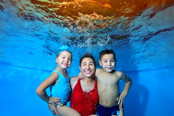 Obraz na płótnie Canvas A beautiful, cheerful family: a mother, a young son and daughter, swimming and posing underwater in the pool. They look at the camera and smile. The concept of a family vacation. Portrait. Close-up.
