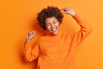 Ecstatic carefree woman with Afro hair dances with arms raised up smiles broadly makes winner gesture celebrates triumph wears orange jumper poses indoor. Joyful dark skinned female moves happily