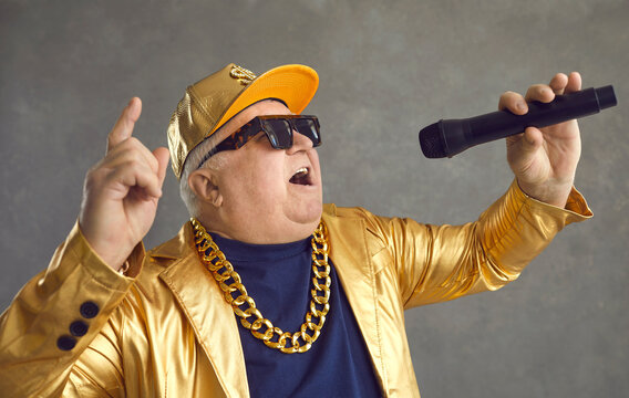 Happy Fat Retired Mature Man Holding Mic, Rapping And Having Fun. Portrait Of Funny Chubby Elderly Pensioner In Baseball Cap, Gold Chain Necklace And Sunglasses With Microphone Singing With Loud Voice